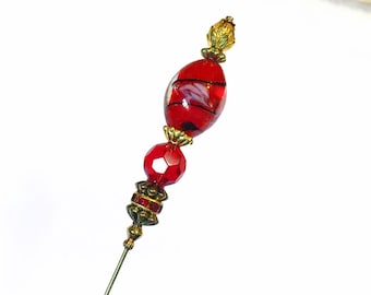 May include: A decorative hat pin with a red glass bead and gold-colored metal accents. The pin features a faceted red bead, a red and black oval bead, and a decorative gold-colored top.