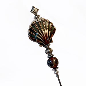 May include: A decorative stick pin featuring a top ornament shaped like a scalloped seashell with a glossy green and brown finish. The pin includes a silver-toned metal finial, a small ribbed silver bead, and a round patterned glass bead below.