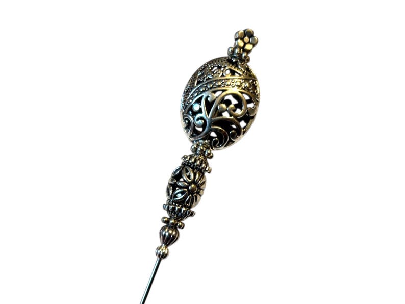 May include: A silver-toned hat pin with an ornate, filigree design. The pin features a decorative oval top with intricate cut-out patterns and a floral-like finial. The stem is slender and pointed, suitable for securing headwear.