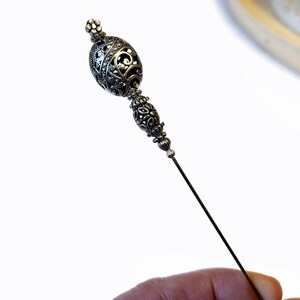 May include: A silver-toned hat pin with an ornate, filigree design. The pin features a decorative, oval-shaped head with intricate patterns and a long, slender pin. The design is detailed and elegant.