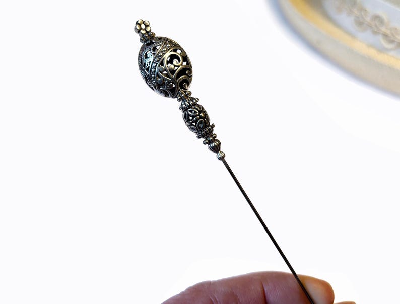 May include: A silver-toned hat pin with an ornate, filigree design. The pin features a decorative, oval-shaped head with intricate patterns and a long, slender pin. The design is detailed and elegant.