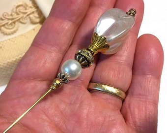 May include: A white pearl and gold tone pin with a large white pearl-like top. The pin is held in a hand.