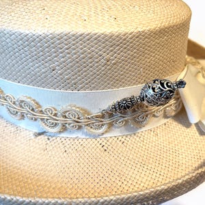 May include: A straw hat with a cream-colored ribbon and decorative trim. The hat features an ornate silver-toned embellishment. The hat's brim is approximately 12 inches in diameter, and the crown is about 5 inches tall.