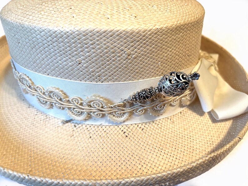 May include: A straw hat with a cream-colored ribbon and decorative trim. The hat features an ornate silver-toned embellishment. The hat's brim is approximately 12 inches in diameter, and the crown is about 5 inches tall.