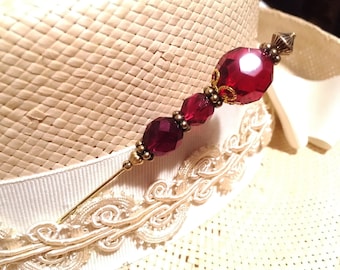 May include: A gold hat pin with a burgundy glass bead and a gold metal top. The pin is on a straw hat with a white ribbon.