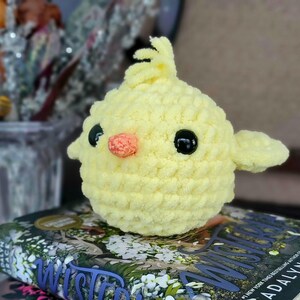 May include: A crocheted yellow chick toy with black eyes and an orange beak. The chick is sitting on a book with the word "Wisteria" visible.