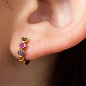 May include: A gold hoop earring with five colorful gemstones: green, pink, blue, and yellow. The earring is worn on an ear, showcasing the vibrant colors of the stones.