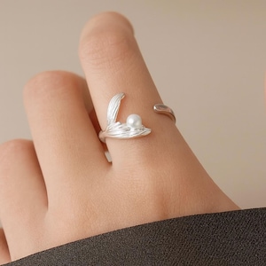 May include: A silver ring with a leaf-like design holding a small, round, white pearl. The ring has an open design for adjustable sizing. The ring is worn on a finger, highlighting its delicate details and elegant simplicity. The ring is made of silver.