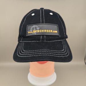 Puede incluir: Gorra de béisbol negra con costuras blancas y visera curva. La gorra presenta un parche rectangular con el texto "www.SPYCHASER.com" en amarillo. La gorra se muestra en una cabeza de maniquí.