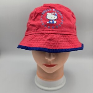 Puede incluir: Un sombrero de pescador rojo con un gráfico de Hello Kitty centrado en la parte delantera. El personaje está rodeado por un borde circular de estrellas azules y el texto HELLO KITTY. El sombrero tiene un borde azul oscuro en la base.