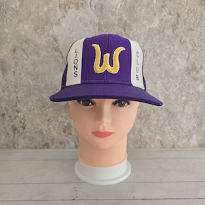 Puede incluir: Una gorra de béisbol morada y blanca con el texto "LIONS CLUB" y un logotipo "W" amarillo. La gorra tiene una visera plana y se muestra en una cabeza de maniquí.