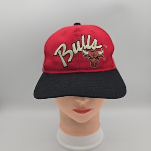 Puede incluir: Una gorra de béisbol roja con visera negra que presenta la palabra Bulls bordada en blanco con un borde negro y un logotipo de cabeza de toro en rojo y blanco. La gorra tiene una corona estructurada con un botón negro en la parte superior.