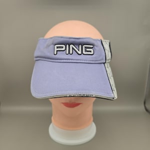 May include: A light purple visor with the word "PING" embroidered in white. The visor has a curved brim and a white and black patterned design on the sides. The text "PLAY YOUR BEST" is printed on the bottom edge.