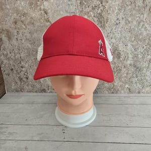 Può includere: Cappellino da baseball rosso e bianco con una parte posteriore in rete bianca e una parte anteriore rossa. Il cappellino ha una "A" bianca ricamata con un alone sul davanti.