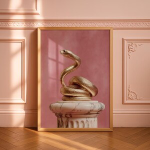 Pode incluir: Uma impressão de arte emoldurada com uma escultura de cobra dourada sobre um pedestal de mármore. A cobra está enrolada e em pé, contra um fundo rosa. A moldura é dourada e a obra de arte é exibida em uma sala com paredes rosa e piso de madeira.