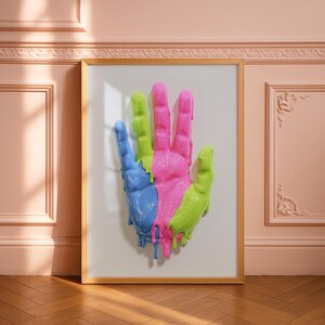 May include: A framed artwork showing a hand sculpture in vibrant colours. The hand is split into blue, pink, and green sections, with a dripping paint effect. The artwork is set against a white backdrop within a gold frame.