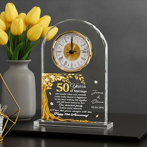 Könnte beinhalten: Eine klare Kristalluhr mit goldenem Zifferblatt und Zeigern. Die Uhr ist mit "50 Jahre Ehe" graviert und enthält Namen und ein Datum. Im Hintergrund steht eine Vase mit gelben Tulpen.