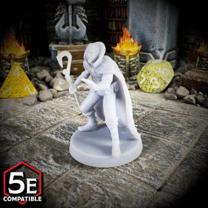 May include: A light gray miniature figure of a robed character holding a staff, posed in a crouched position. The figure is on a round base, with the text "5E COMPATIBLE" in a red and white hexagon. The figure is likely for tabletop gaming.