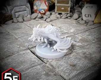 May include: A white dragon miniature figurine with detailed wings and a textured body, posed on a round base. The figurine is set on a stone tile surface, with a 5E Compatible logo in the corner. Other tabletop gaming elements are in the background.