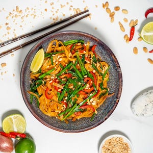 Peut inclure: Un bol de nouilles Pad Thai avec des cacahuètes, des poivrons rouges et des oignons verts. Les nouilles sont sur une assiette bleu foncé avec un fond blanc. Il y a des baguettes, des citrons verts et des cacahuètes éparpillées autour de l'assiette.