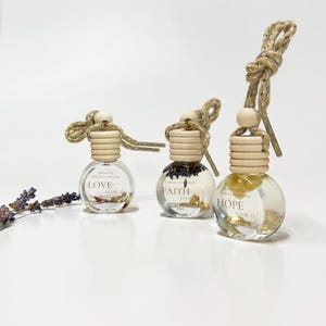 Puede incluir: Tres pequeñas botellas de vidrio con tapones de corcho y una cuerda dorada cuelgan de una cuenta de madera. Cada botella contiene flores secas y escamas doradas. Las botellas están etiquetadas con "LOVE", "FAITH" y "HOPE".