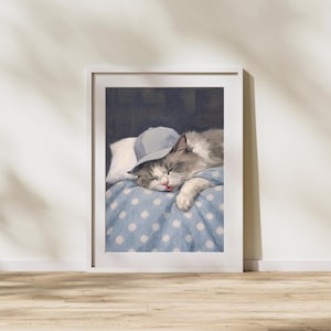 Puede incluir: Una impresión enmarcada de un gato durmiendo con una gorra de béisbol azul claro. El gato está descansando sobre una manta azul con lunares y una almohada blanca. El pelaje del gato es gris y blanco, y tiene la lengua fuera.