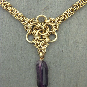 Puede incluir: Un collar de cadena de oro con un colgante que presenta un dise&ntilde;o de nudo tejido y una piedra preciosa p&uacute;rpura.