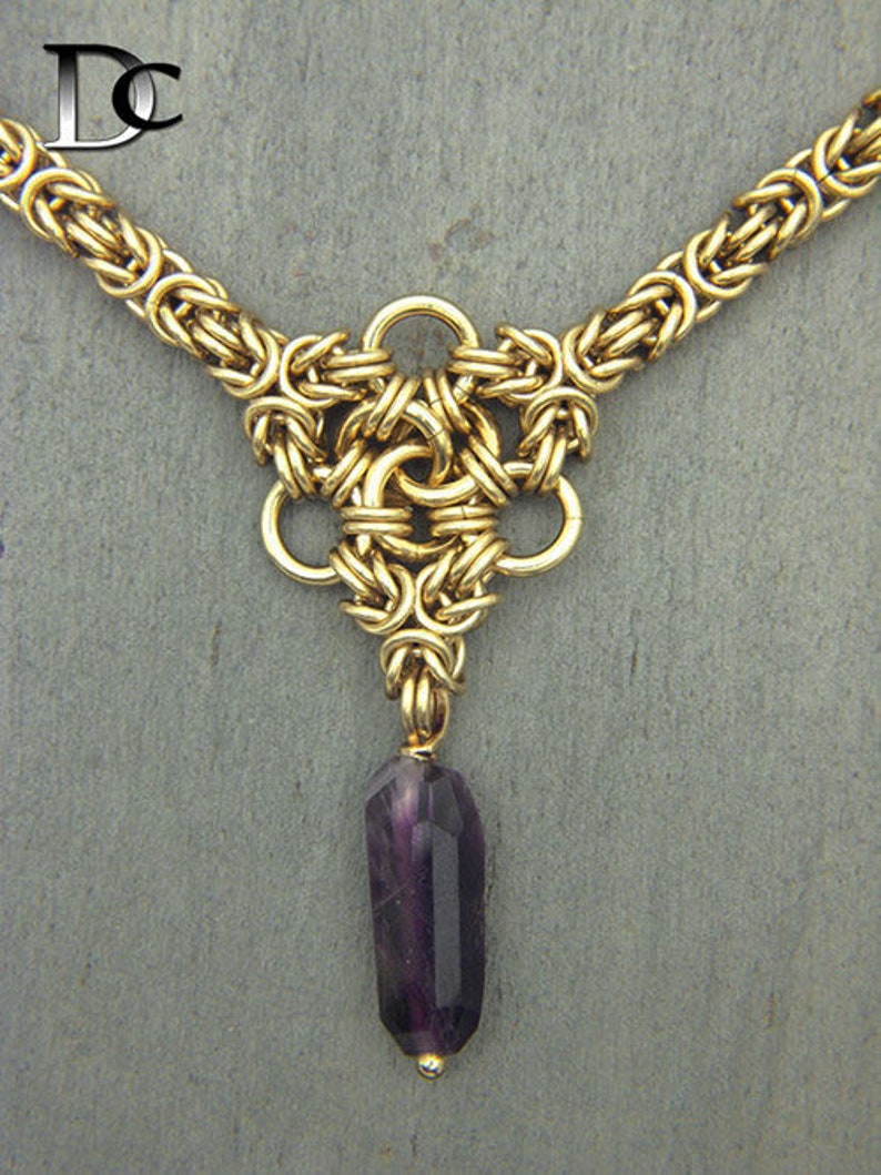 Puede incluir: Un collar de cadena de oro con un colgante que presenta un dise&ntilde;o de nudo tejido y una piedra preciosa p&uacute;rpura.