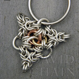 Puede incluir: Un llavero de plata con un dije triangular hecho de anillos de metal entrelazados. El dije tiene un anillo central de cobre y lat&oacute;n.