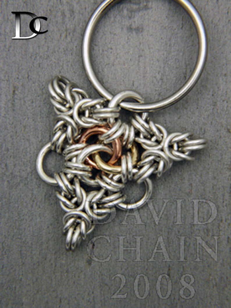 Puede incluir: Un llavero de plata con un dije triangular hecho de anillos de metal entrelazados. El dije tiene un anillo central de cobre y lat&oacute;n.