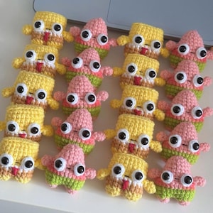 Peut inclure: Une collection de petites figurines crochetées à la main disposées en rangées. Les figurines alternent entre des personnages jaunes de forme carrée et des personnages roses en forme d'étoile, avec de grands yeux en plastique noir et blanc.