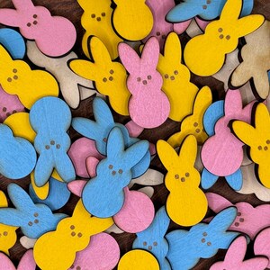 May include: A collection of colorful wooden bunny shapes in pink, blue, and yellow. The bunnies have a simple design with two eyes and a mouth.