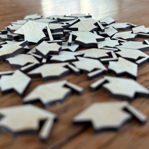 May include: A pile of wooden graduation cap shapes, painted white with black outlines. The caps are scattered on a wooden surface.