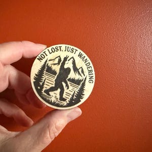 May include: A round wooden coaster with a Bigfoot design. The image features a silhouette of Bigfoot with the text "NOT LOST, JUST WANDERING" around the edge. The design includes trees and mountains.