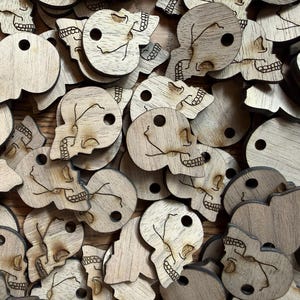 May include: A pile of small, wooden skull-shaped cutouts. Each cutout has a hole at the top and detailed etched features, including eye sockets, a nose, and a grinning mouth. The wood grain varies in color from light to dark.
