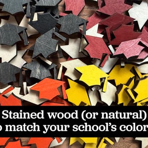 May include: A collection of wooden graduation caps in various colors, including black, red, yellow, and natural wood tones. The caps are scattered on a wooden surface. Text on the image reads: "Stained wood (or natural) to match your school's colors!"