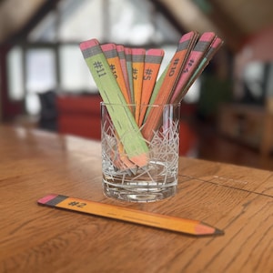 May include: A set of colorful wooden pencils with numbers on them, arranged in a glass. The pencils are in a glass on a wooden table. One pencil is lying on the table, labeled #2.