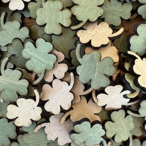 May include: A pile of wooden four-leaf clover charms in green and natural wood tones. The charms are scattered on a wooden surface.
