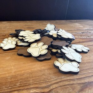 May include: A pile of wooden paw print cutouts. The paw prints have a light-colored center with a dark brown outline and base. The cutouts are scattered on a wooden surface.