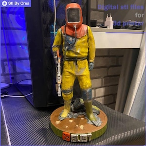 Puede incluir: Una figura impresa en 3D de un personaje del videojuego Rust. El personaje lleva un traje de protección amarillo con un casco rojo y sostiene un arma. La figura está de pie sobre una base con la palabra "Rust" impresa en ella.