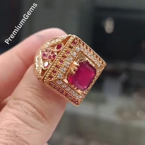 Op de afbeelding: Een goudkleurige ring met een grote, vierkant geslepen donkerrode middensteen, omringd door kleinere witte en rode accentstenen in een geometrische zetting. De metalen band vertoont ingewikkelde filigraanpatronen en de tekst PremiumGems is zichtbaar.