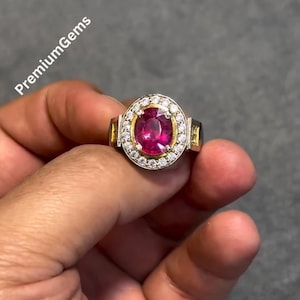 Op de afbeelding: Een gouden ring met een grote ovale robijn edelsteen in het midden, omringd door een halo van kleine, heldere edelstenen. De ring heeft een rechthoekig ontwerp aan de zijkanten. De tekst "PremiumGems" is linksboven zichtbaar.