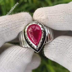May include: A silver ring featuring a large, teardrop-shaped, deep red gemstone. The gemstone is set in a black border, and the ring has a ribbed band. The ring is displayed against a white background.