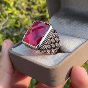 May include: A silver ring with a large, square-cut, ruby-red gemstone. The ring's band features a detailed, textured design with small, red accent stones. The ring is presented in a gray jewelry box, showcasing its intricate craftsmanship.