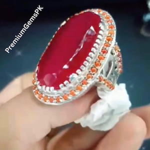 May include: A large, oval-shaped ring with a vibrant red gemstone. The ring features a silver band with a double row of orange and silver accents around the gemstone. The text "PremiumGemsPK" is visible in the upper left corner.