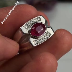 May include: A silver ring featuring a large, oval-cut ruby gemstone at the center. The ring is accented with small, clear gemstones on either side of the ruby. The ring is held in a hand.