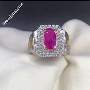May include: A silver and gold ring featuring a large, oval-cut ruby gemstone. The ruby is surrounded by a halo of small, clear gemstones. The ring has a rectangular face and a textured band. The text "Premium Gems" is visible.