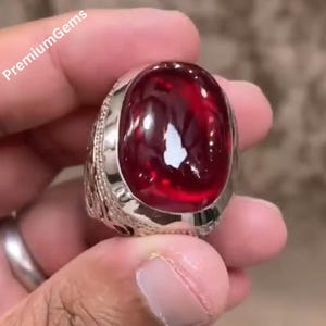 May include: A silver ring with an oval, deep red gemstone. The ring's band features intricate detailing. The text "PremiumGems" is visible in the upper left corner.