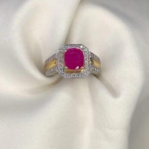 May include: A silver ring featuring a large, square, magenta gemstone. The gemstone is encircled by small, clear stones. The band has gold-coloured accents. The ring is set against a white background.