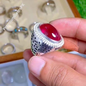 May include: A silver ring with a large, oval-shaped, deep red gemstone. The ring has a detailed, openwork design on the band and a textured border around the stone. The text "PremiumGemsPK" is visible in the background.